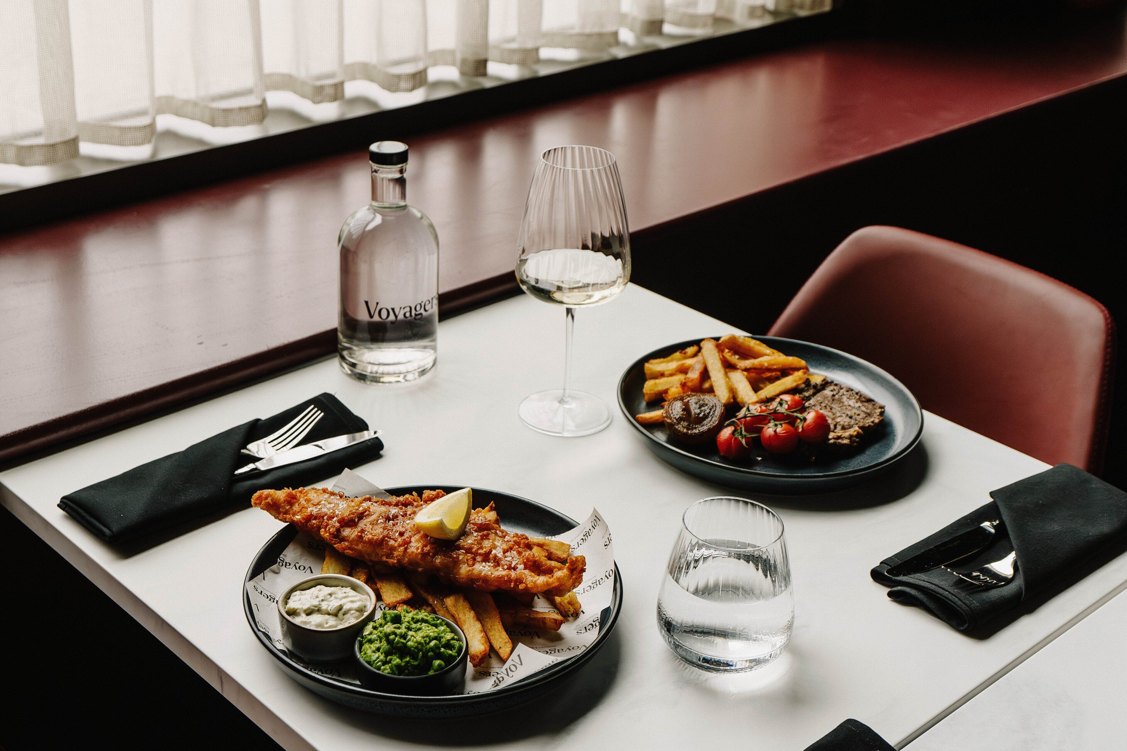 A dining table with a meal consisting of fish and chips, a bottle of water, a glass of wine, and a plate of fries with tomatoes and a slice of bread.