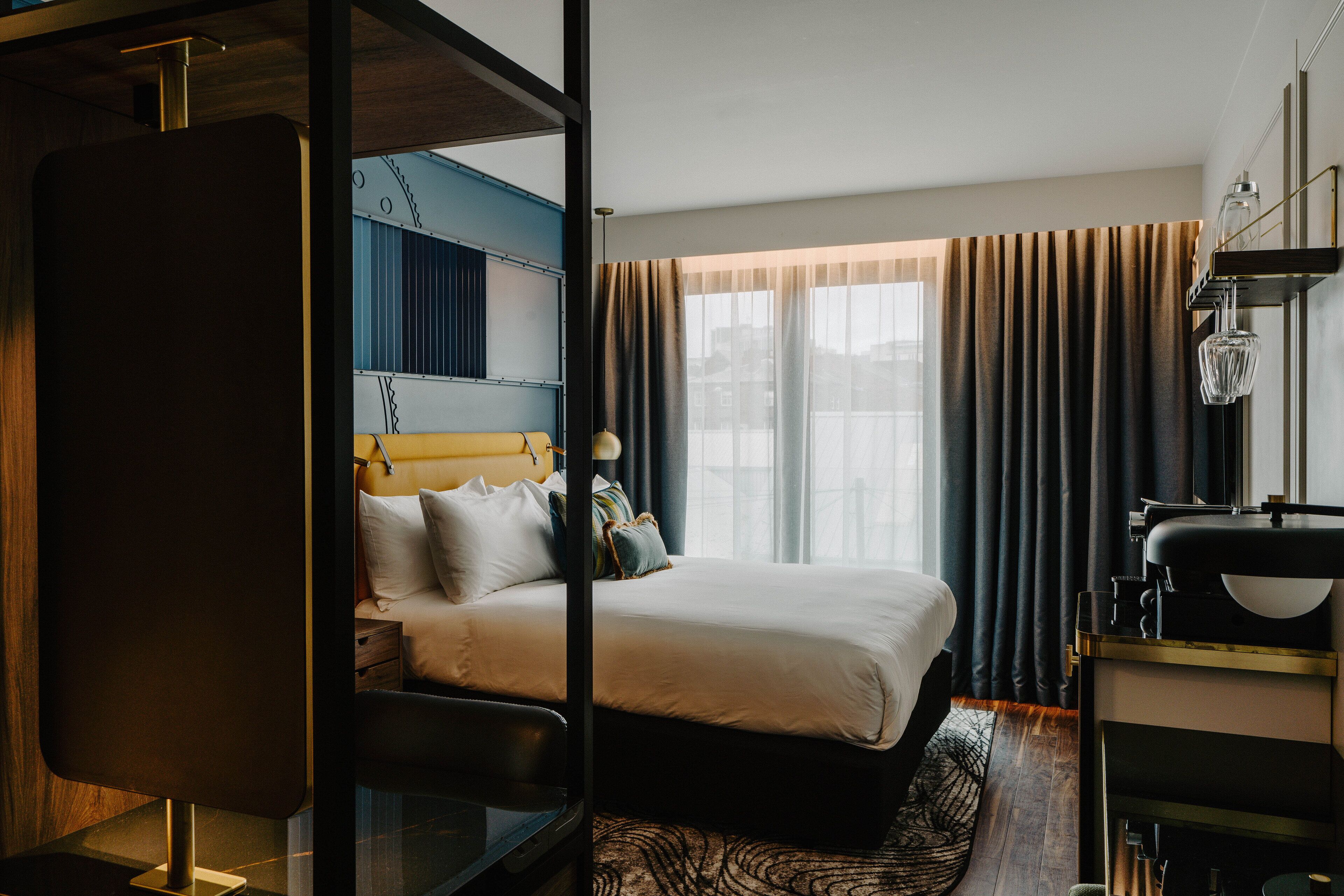 A modern hotel room with a large bed, a desk with a lamp, and a view of the city through large windows.