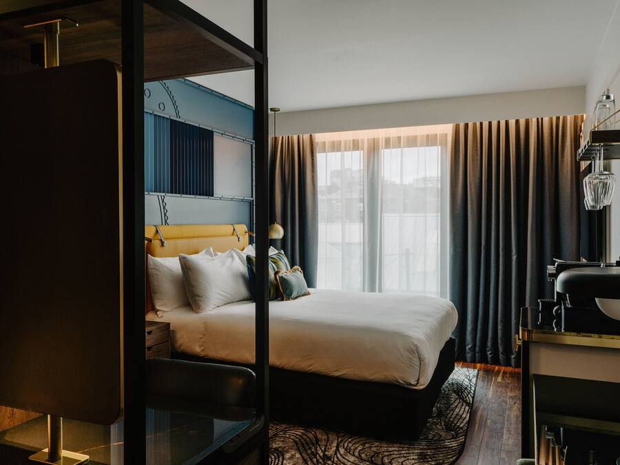 Ein modernes Hotelzimmer mit einem großen Bett, einem Schreibtisch mit Lampe und Blick auf die Stadt durch große Fenster.