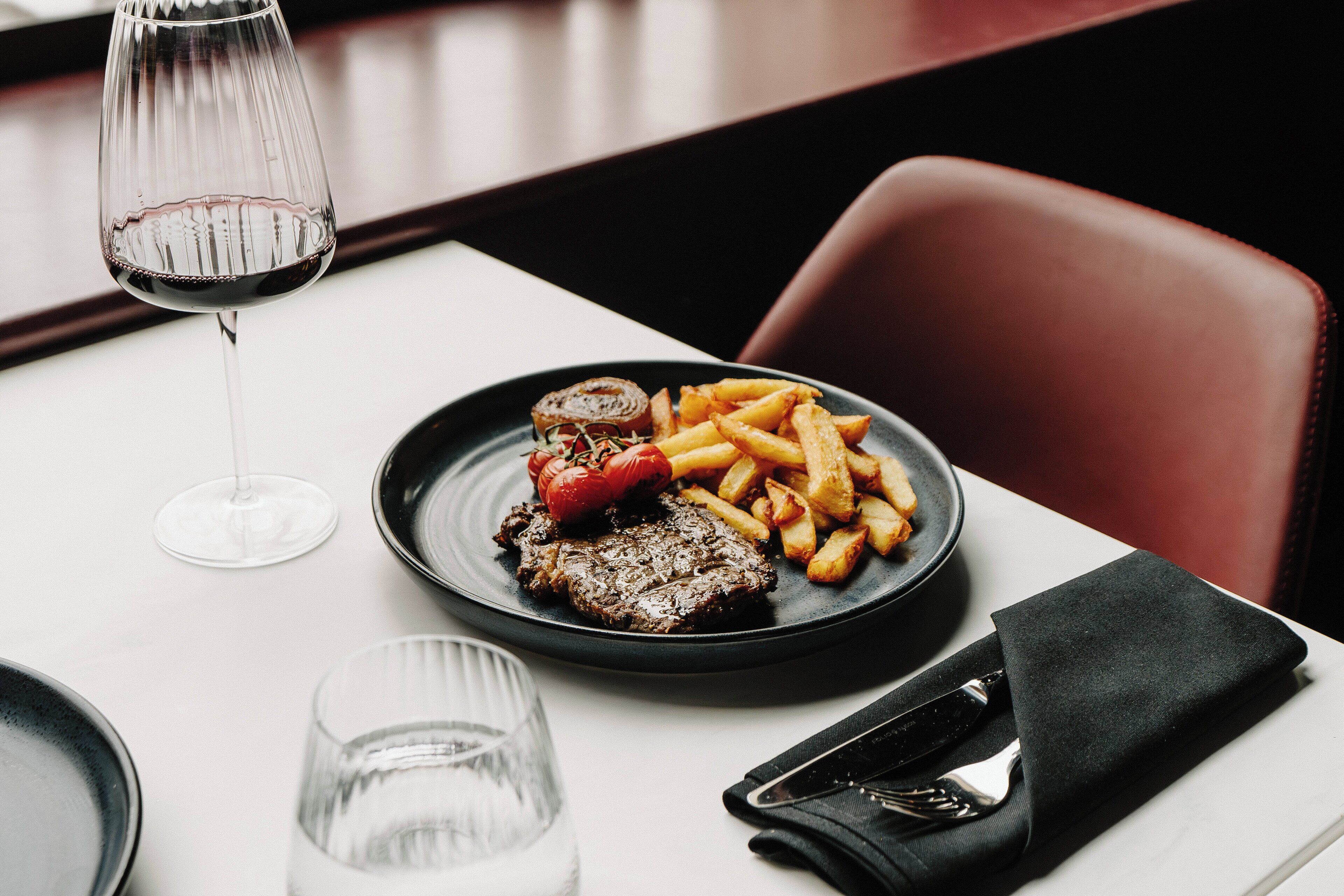 Een eettafel met een steak, friet en tomaat, een wijnglas en bestek.