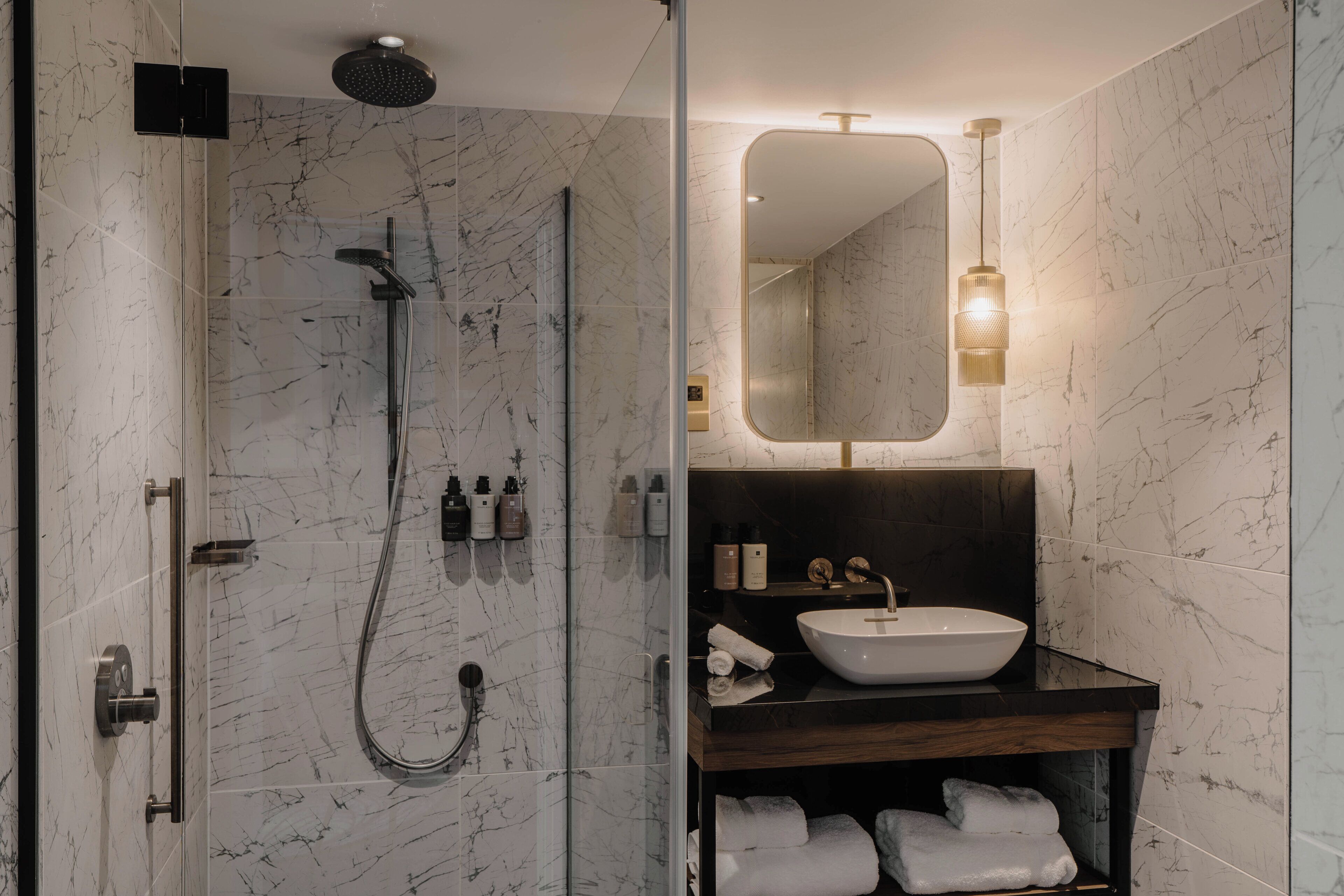 Une salle de bain moderne avec une douche à parois de verre, un meuble-lavabo en bois avec un évier blanc et un miroir surmonté d’un luminaire.