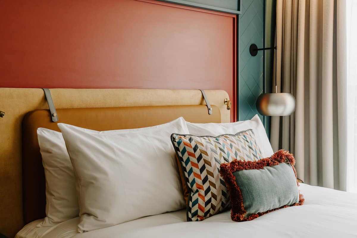 A neatly made bed with white and patterned pillows, a headboard with a strap, and a lamp on the side.