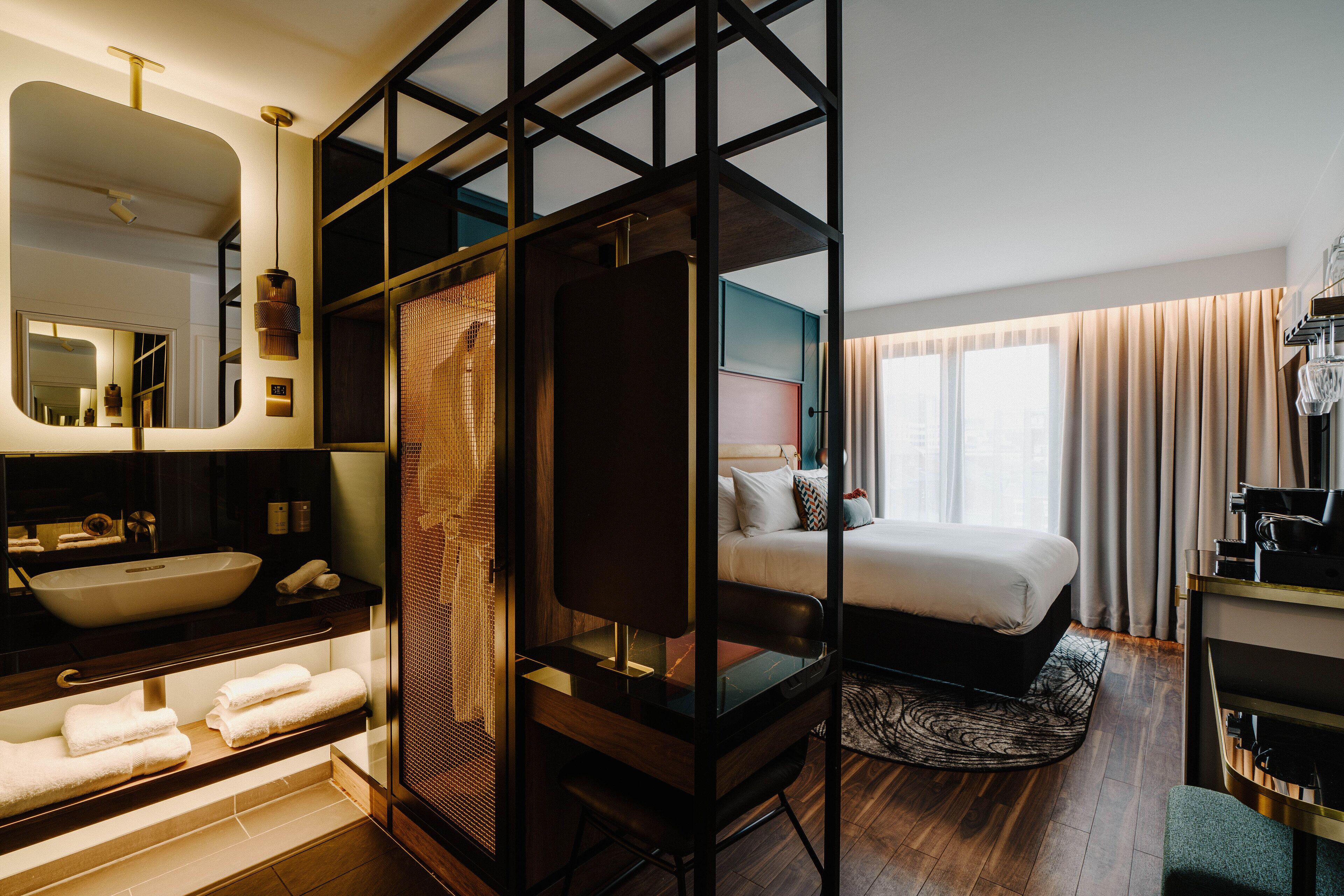 A modern hotel room with a large bed, a separate bathroom with a large mirror, and a view of the city through the window.