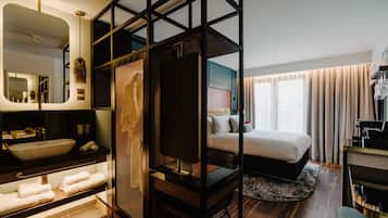 Una habitación de hotel moderna con una cama grande, un baño independiente con un espejo amplio y vistas a la ciudad a través de la ventana.
