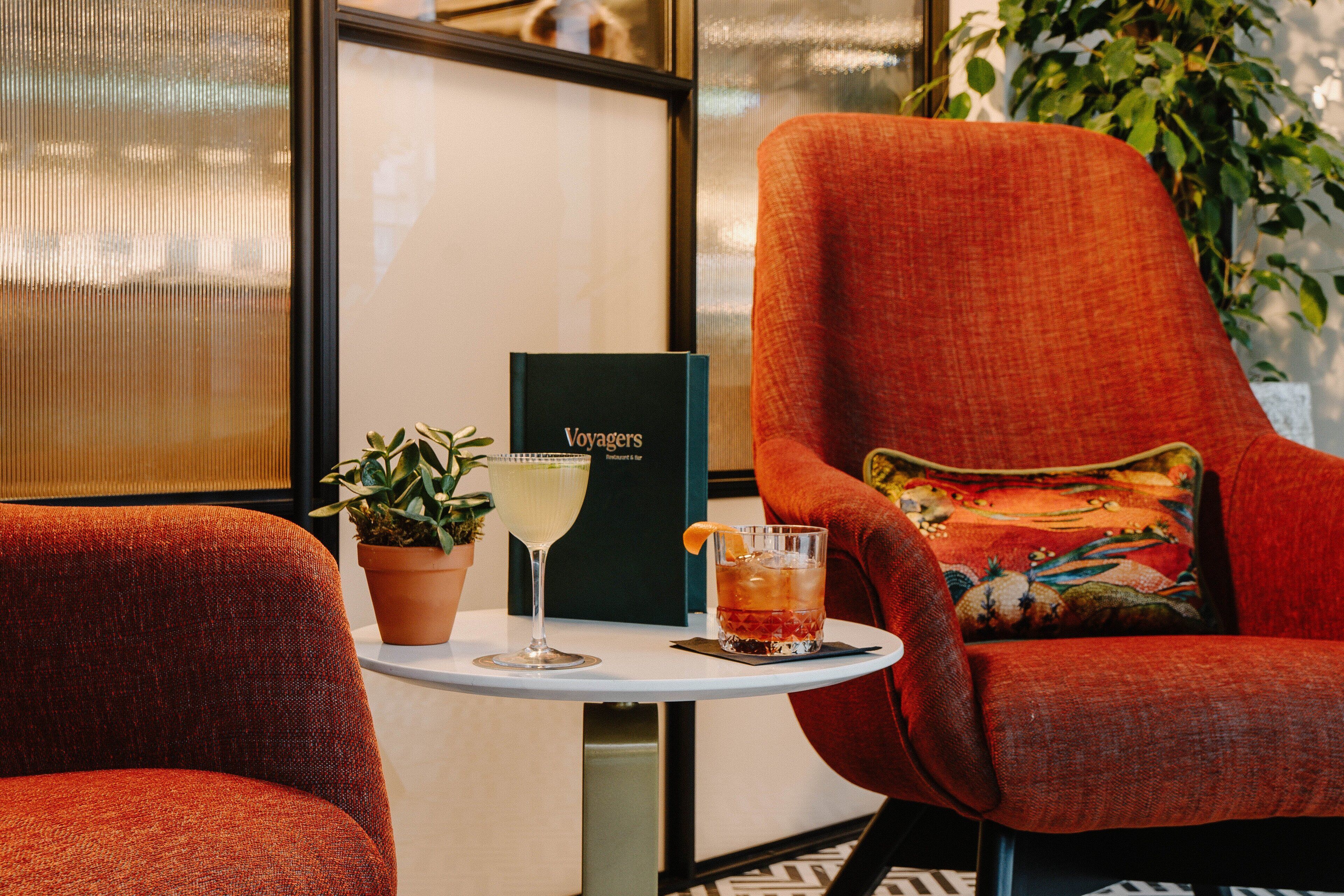 Un fauteuil rouge recouvert de tissu, agrémenté d’un coussin coloré, une petite table sur laquelle est posée une plante en pot, un verre à cocktail et un menu intitulé « Voyagers ».
