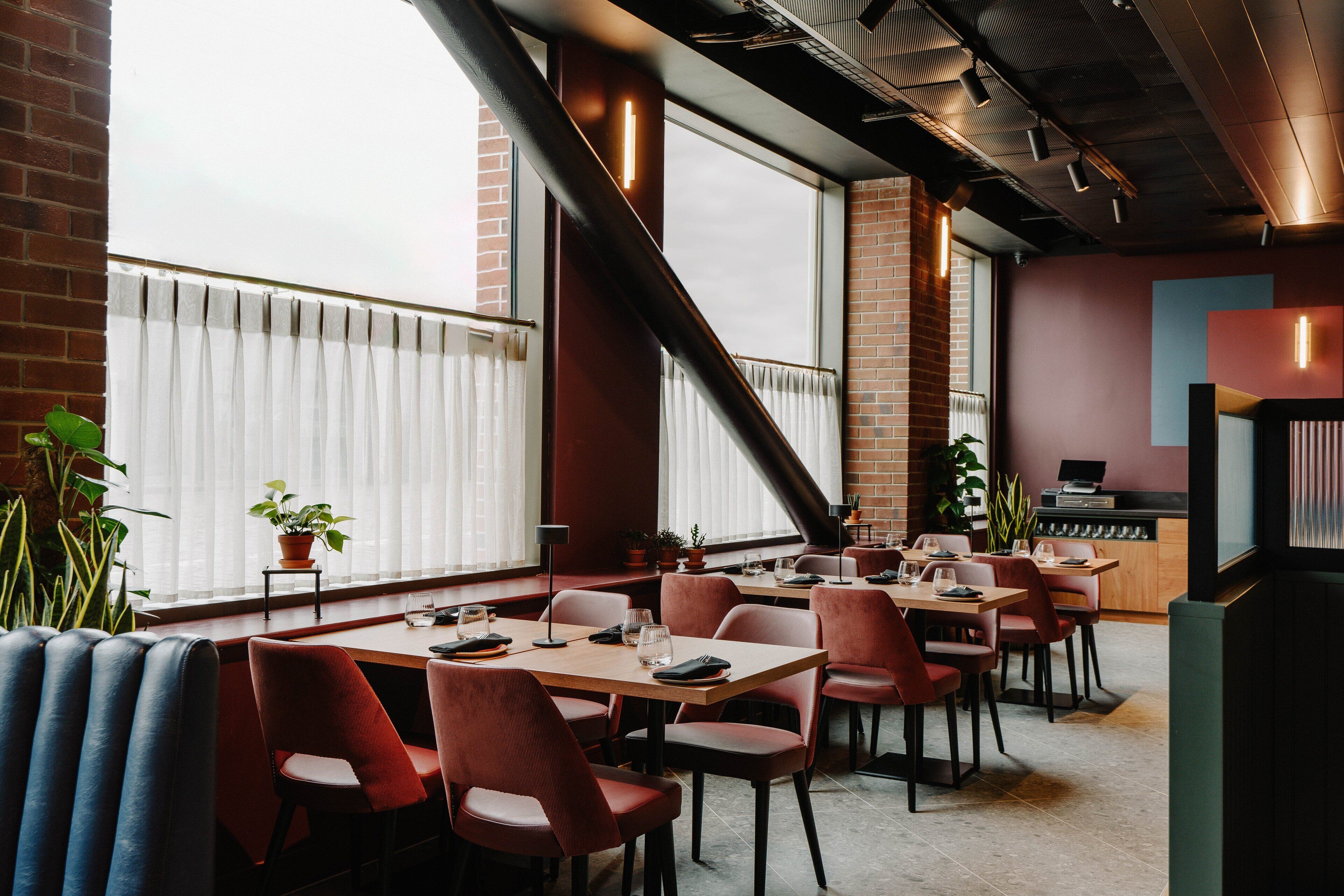 A modern restaurant with brick walls, large windows, and red chairs. Tables are set with glasses and cutlery.