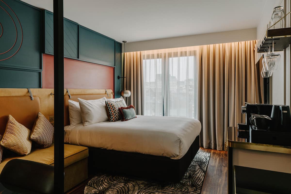 A hotel room with a large bed, a desk with a coffee machine, and a view of the city through sheer curtains.