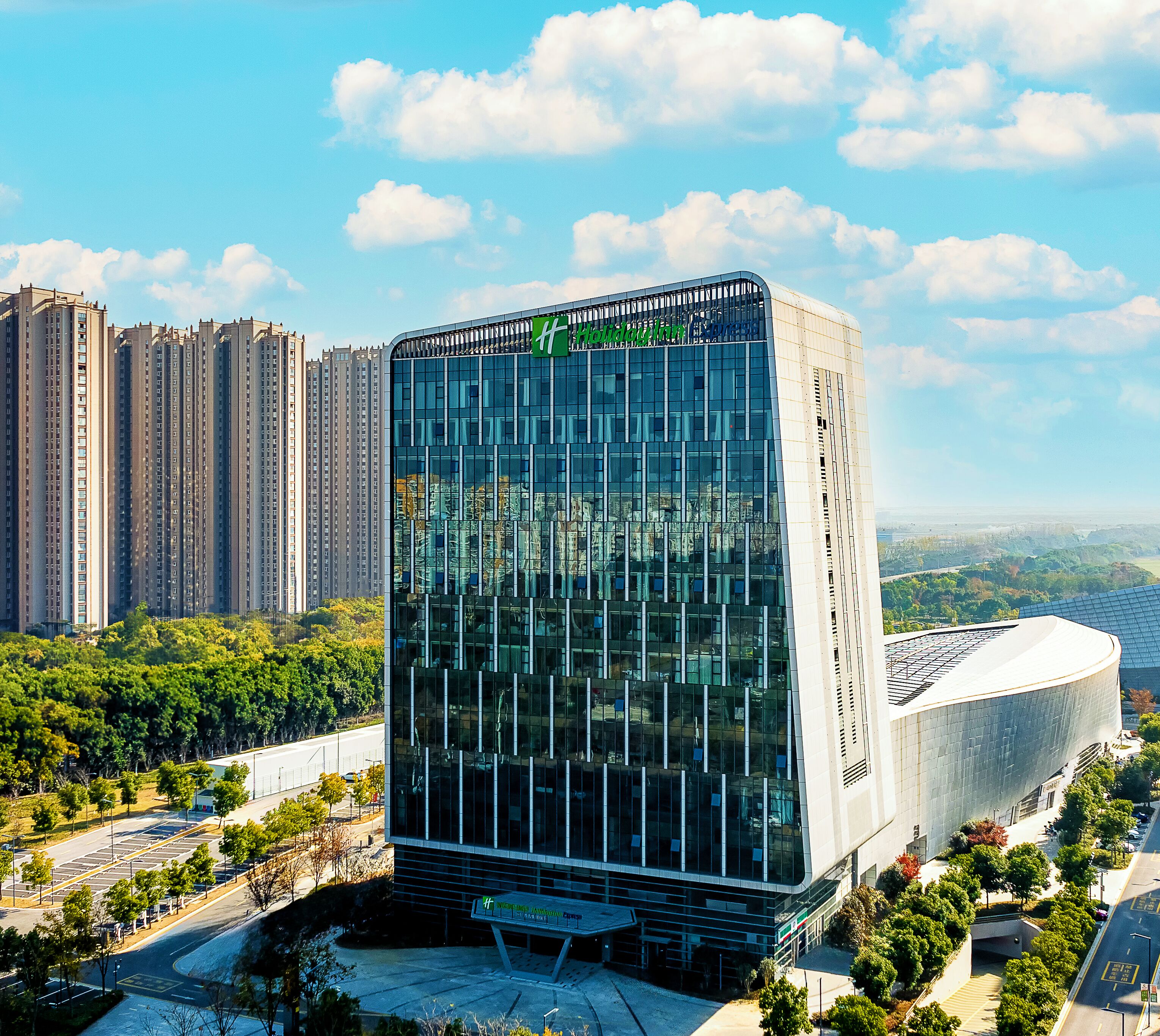 A modern hotel with a glass facade, surrounded by high-rise buildings and greenery.