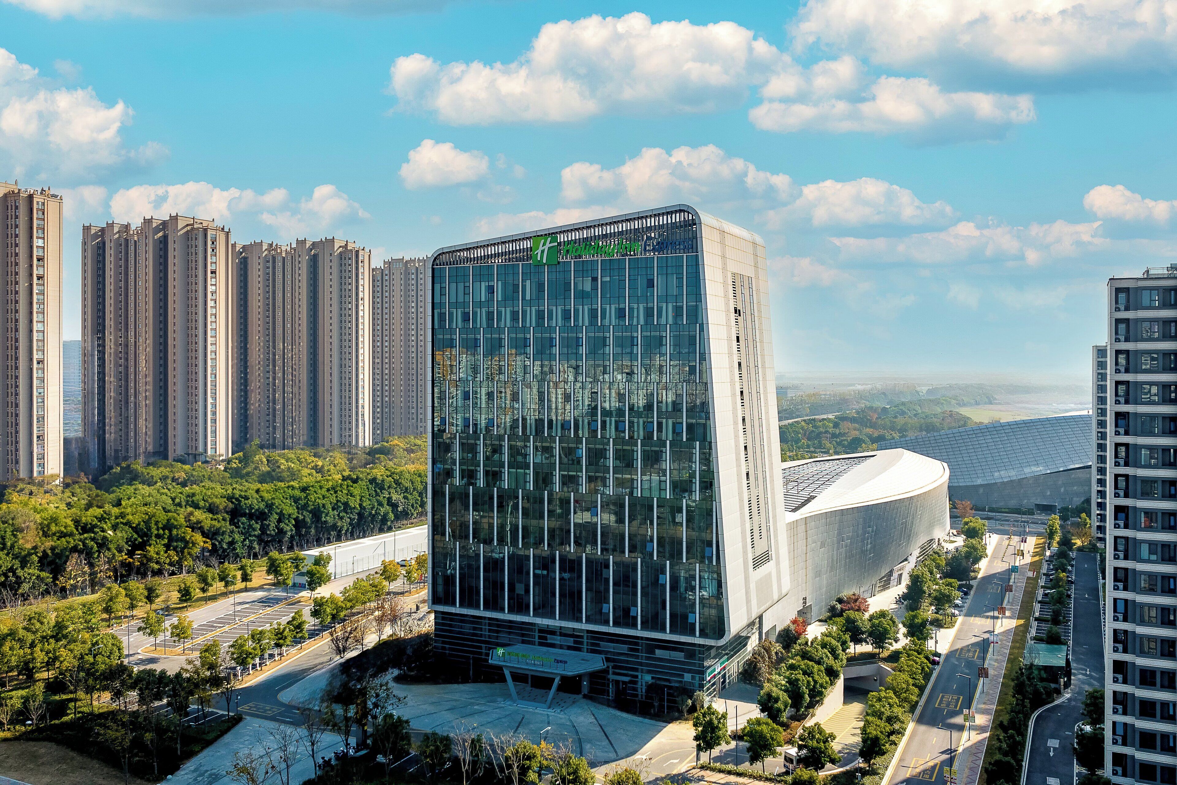 A modern hotel with a glass facade, surrounded by high-rise buildings and a landscaped area.