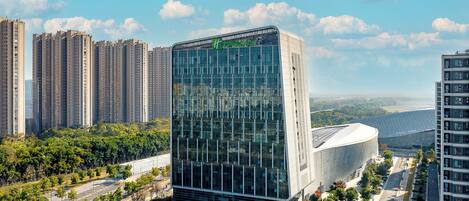 A modern hotel with a glass facade, surrounded by high-rise buildings and a landscaped area.