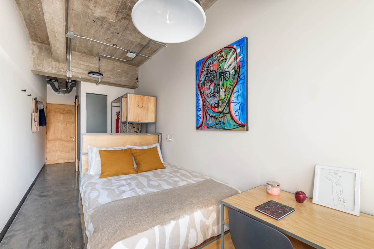 Une chambre moderne avec un lit, un bureau, une chaise, une lampe, un tableau et une porte en bois.