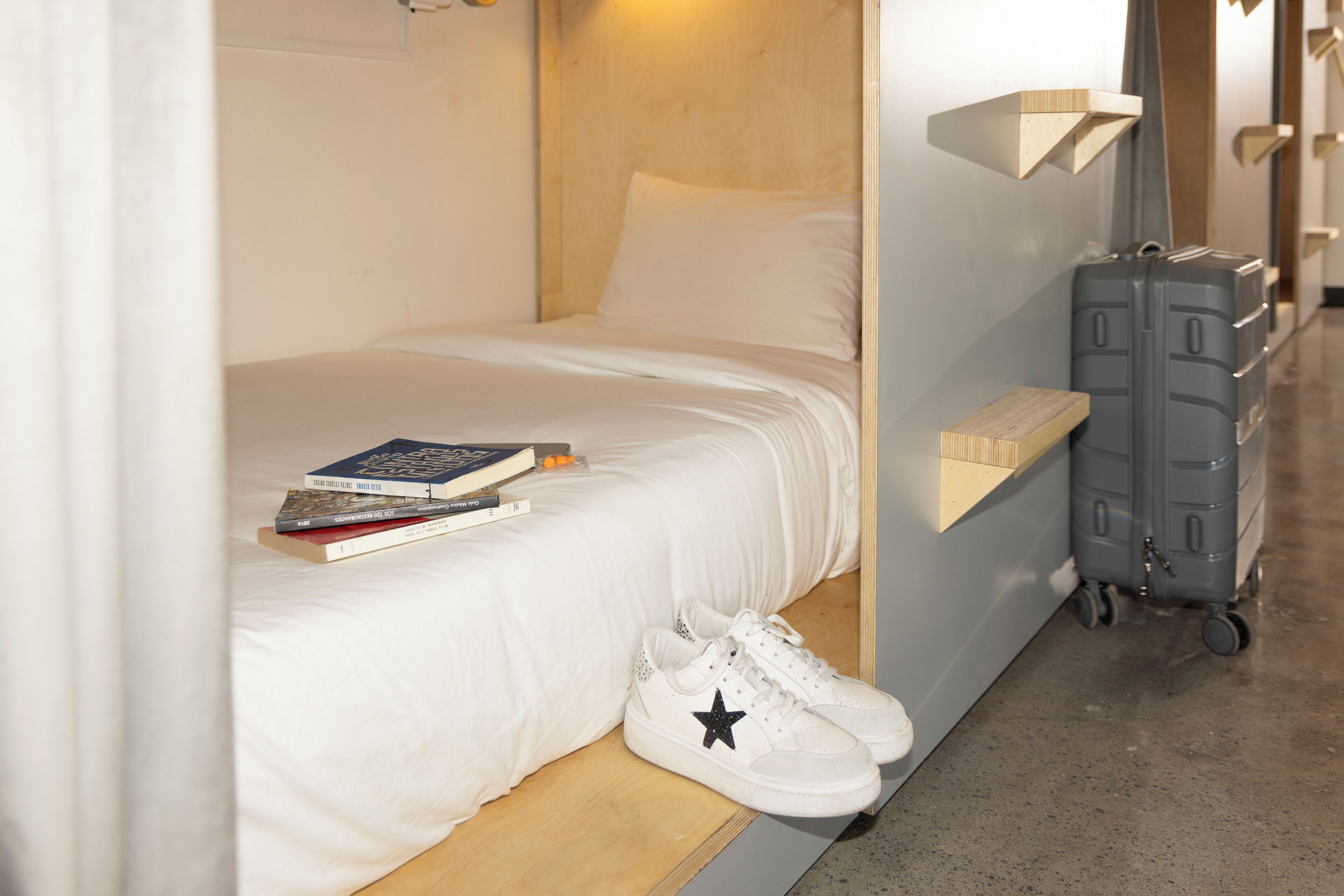 Une chambre d’hôtel de taille réduite, comprenant un lit simple, une petite table de chevet, une valise et une étagère fixée au mur.