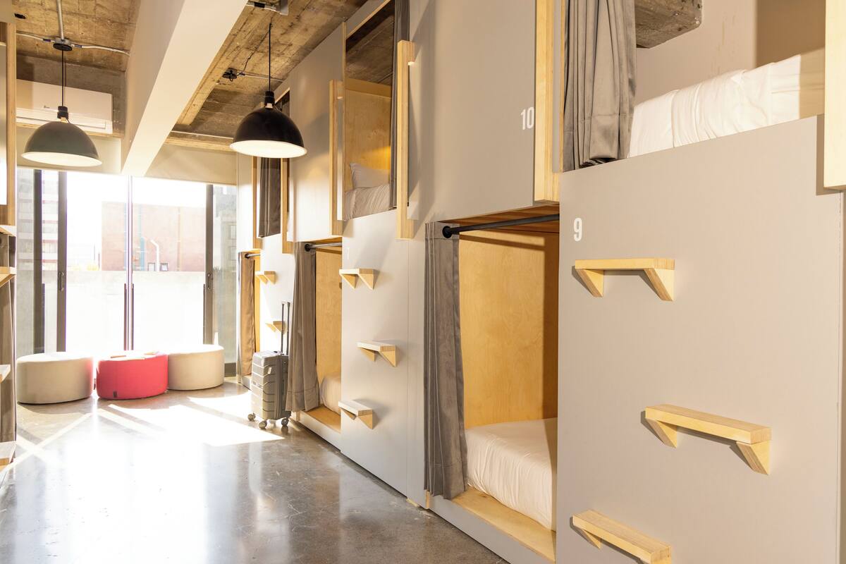 Une chambre d’hôtel moderne équipée de lits superposés, de meubles de rangement en bois et d’une grande fenêtre laissant entrer la lumière naturelle.