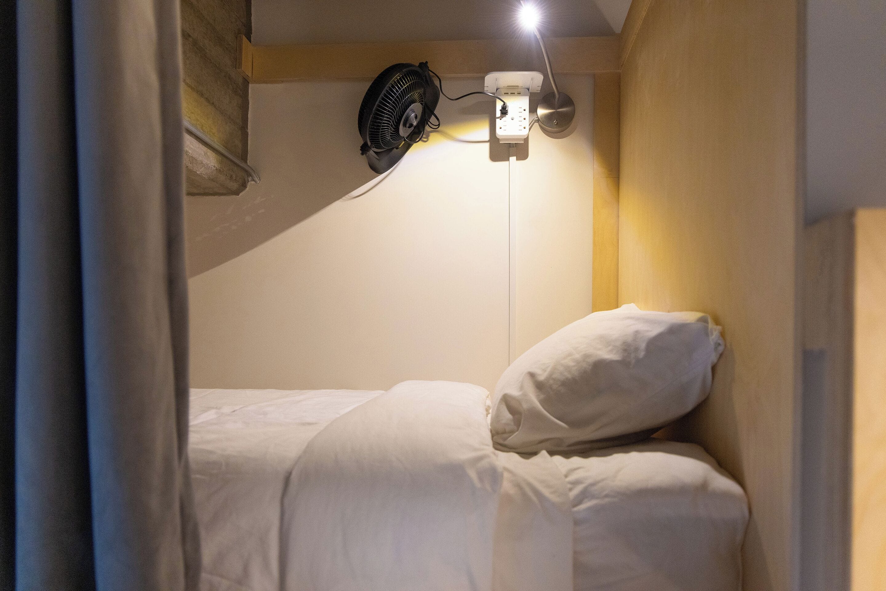 Uma cama de solteiro com roupa de cama branca, um ventilador fixado na parede e uma luminária.