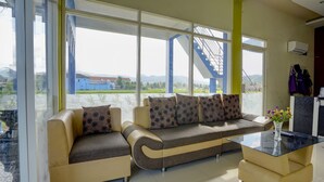 A modern living room with a beige sofa, a glass coffee table, and large windows offering a view of a blue staircase and greenery outside. - OYO 92907 Miranda Hotel Syariah (Gresik)