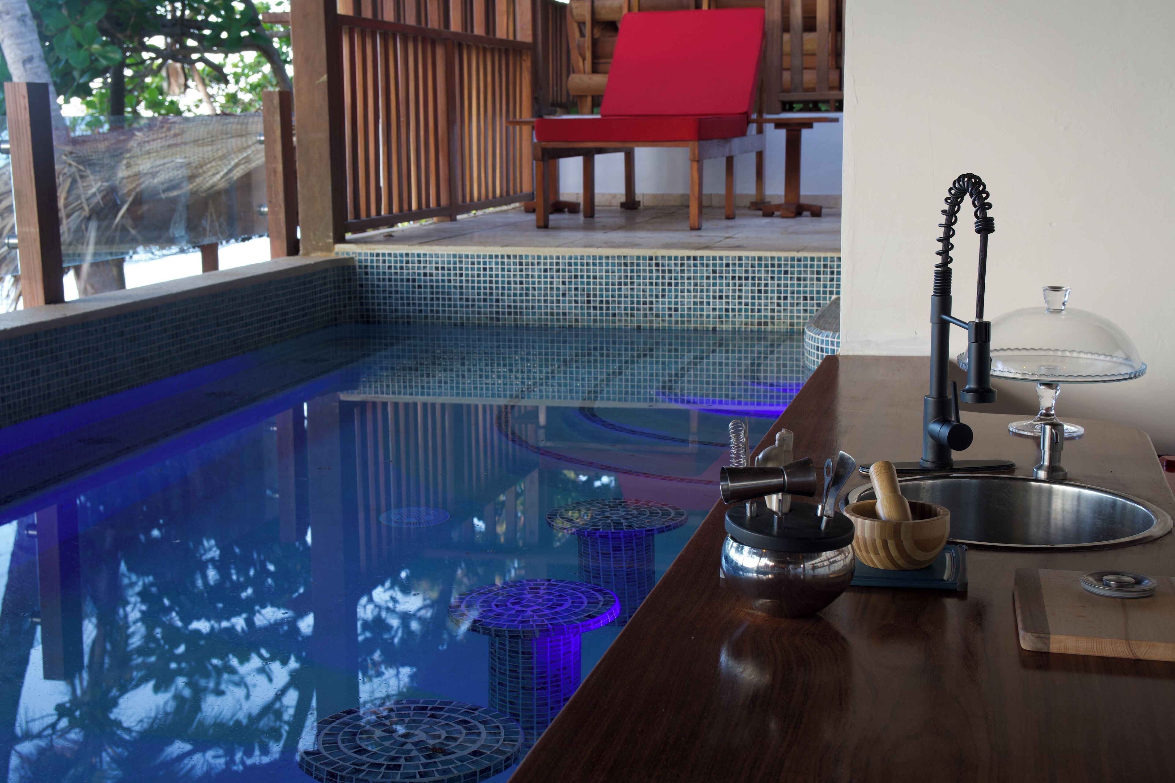 Una piscina con una zona de bar de madera, una silla roja y un escurreplatos con cubierta de vidrio.