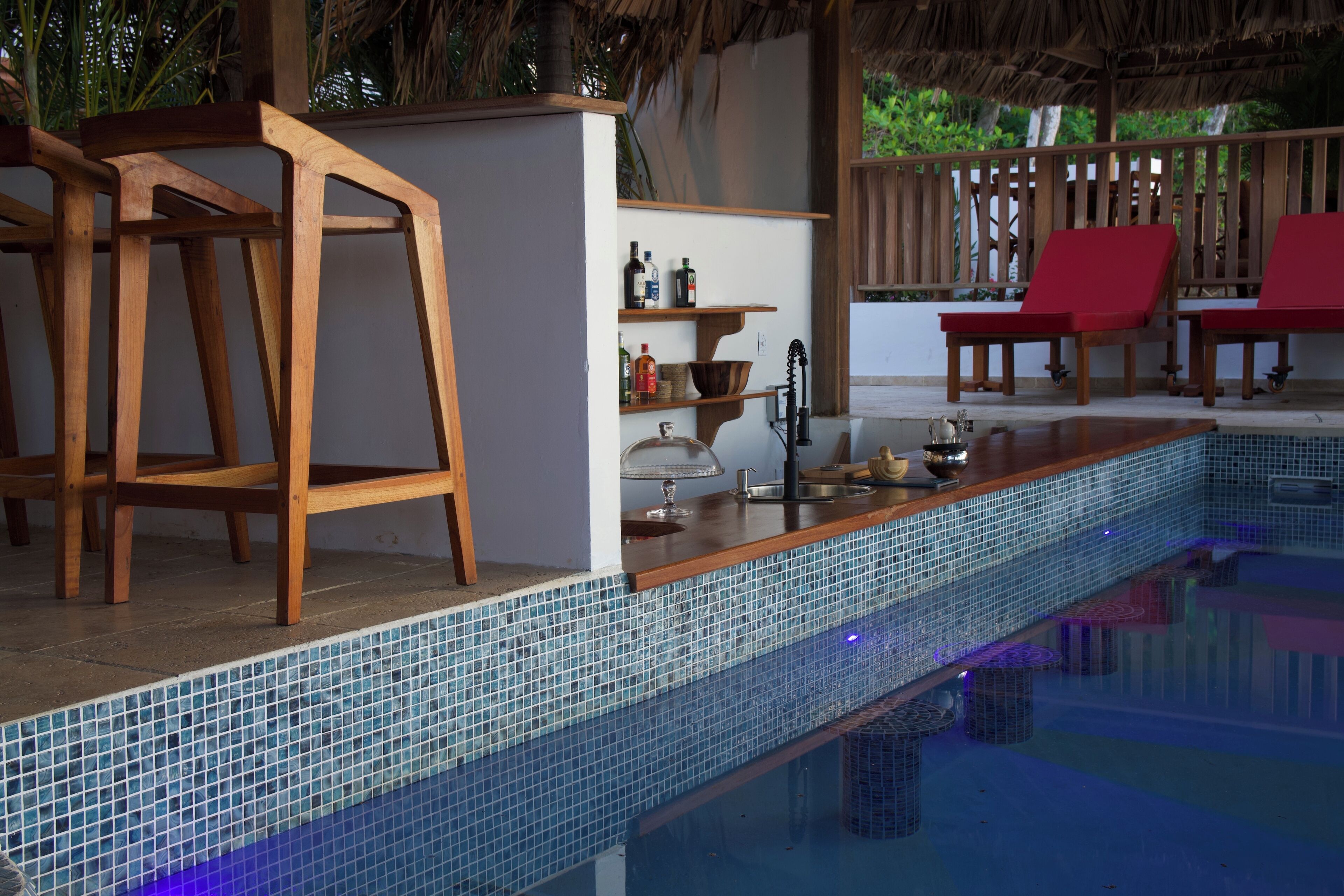 A poolside area with wooden chairs, a bar, and a shelf with bottles and a bowl.