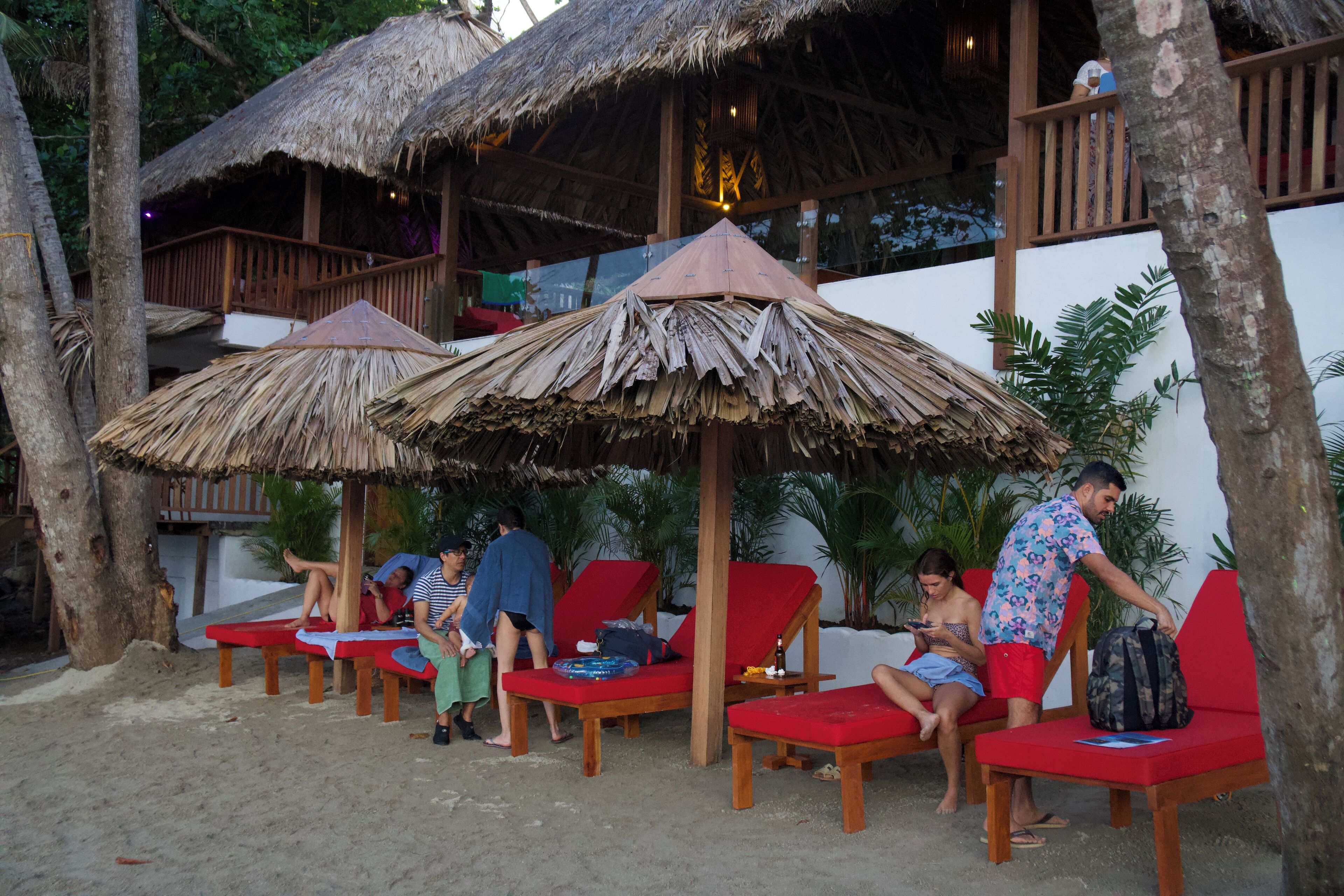 Un salón junto a la playa con sillas de madera y sombrillas de paja, rodeado de palmeras y un edificio blanco.