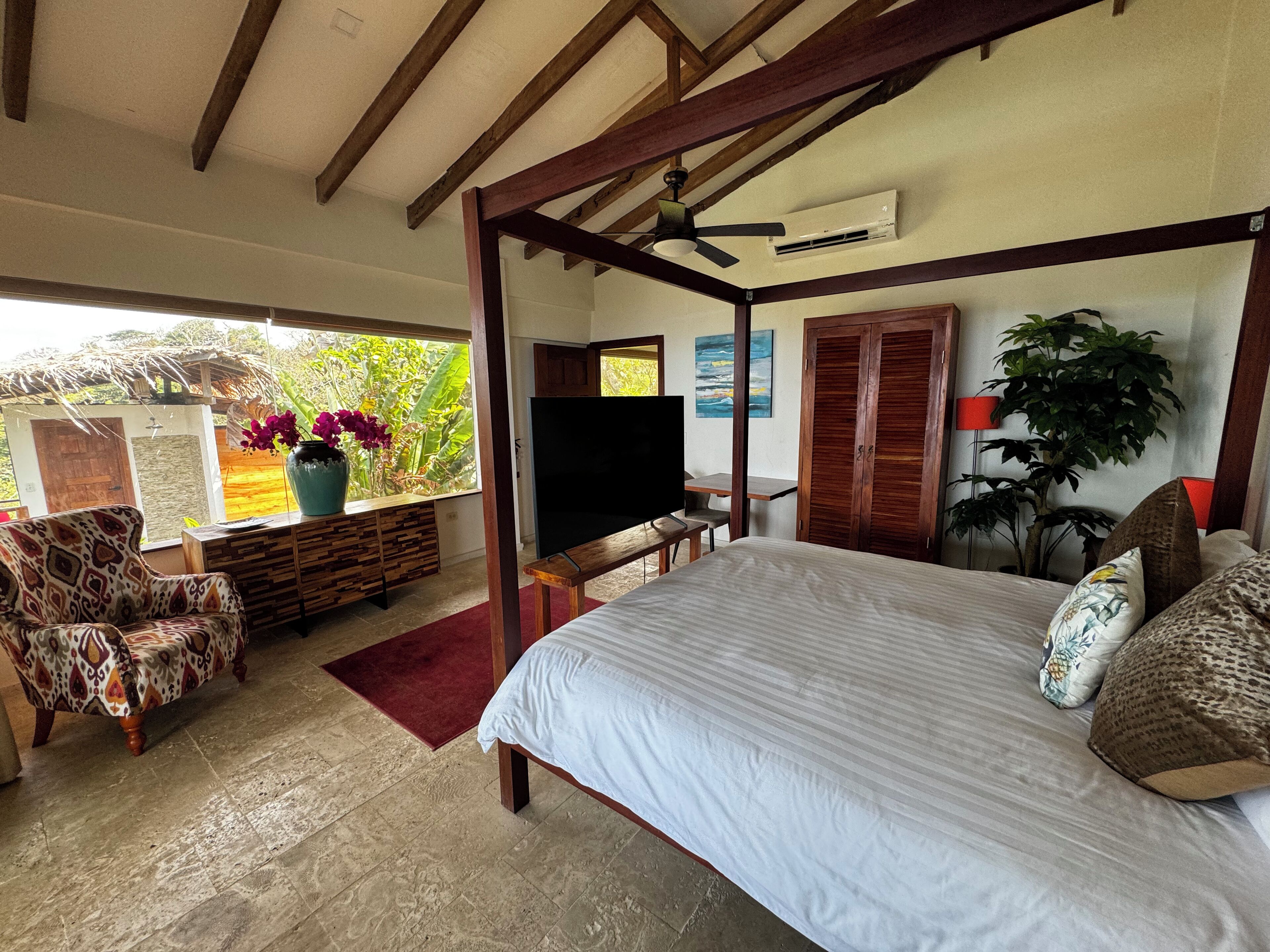 Un dormitorio con cama con dosel, televisión, escritorio y ventana con vista a la vegetación.
