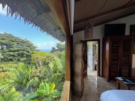 Quarto com uma grande janela que dá para um jardim exuberante e vista para o oceano. Há uma escultura de bambu, uma porta de madeira e uma cama com lençol branco.