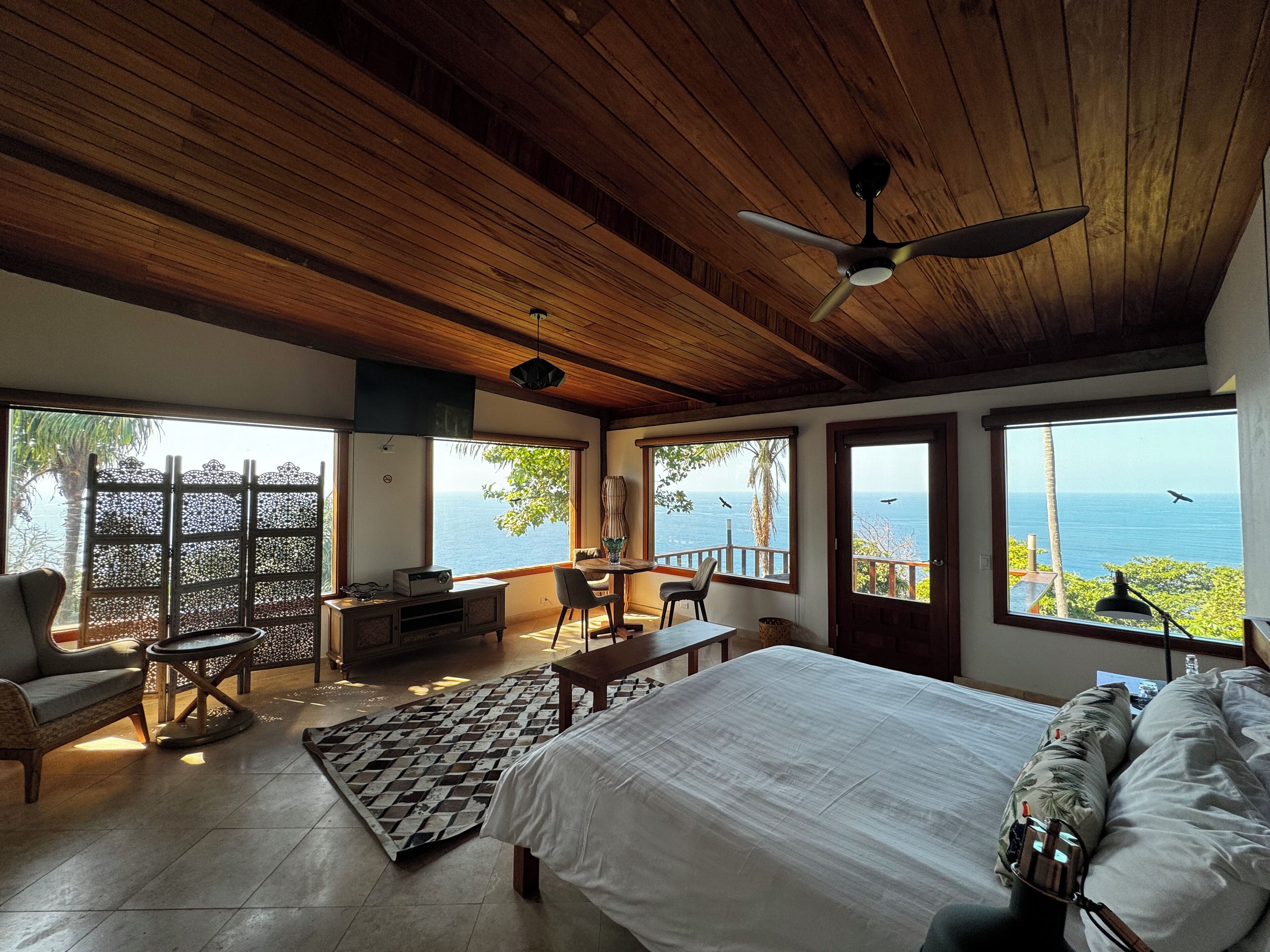 A bedroom with a large bed, wooden ceiling, and multiple windows offering ocean views.