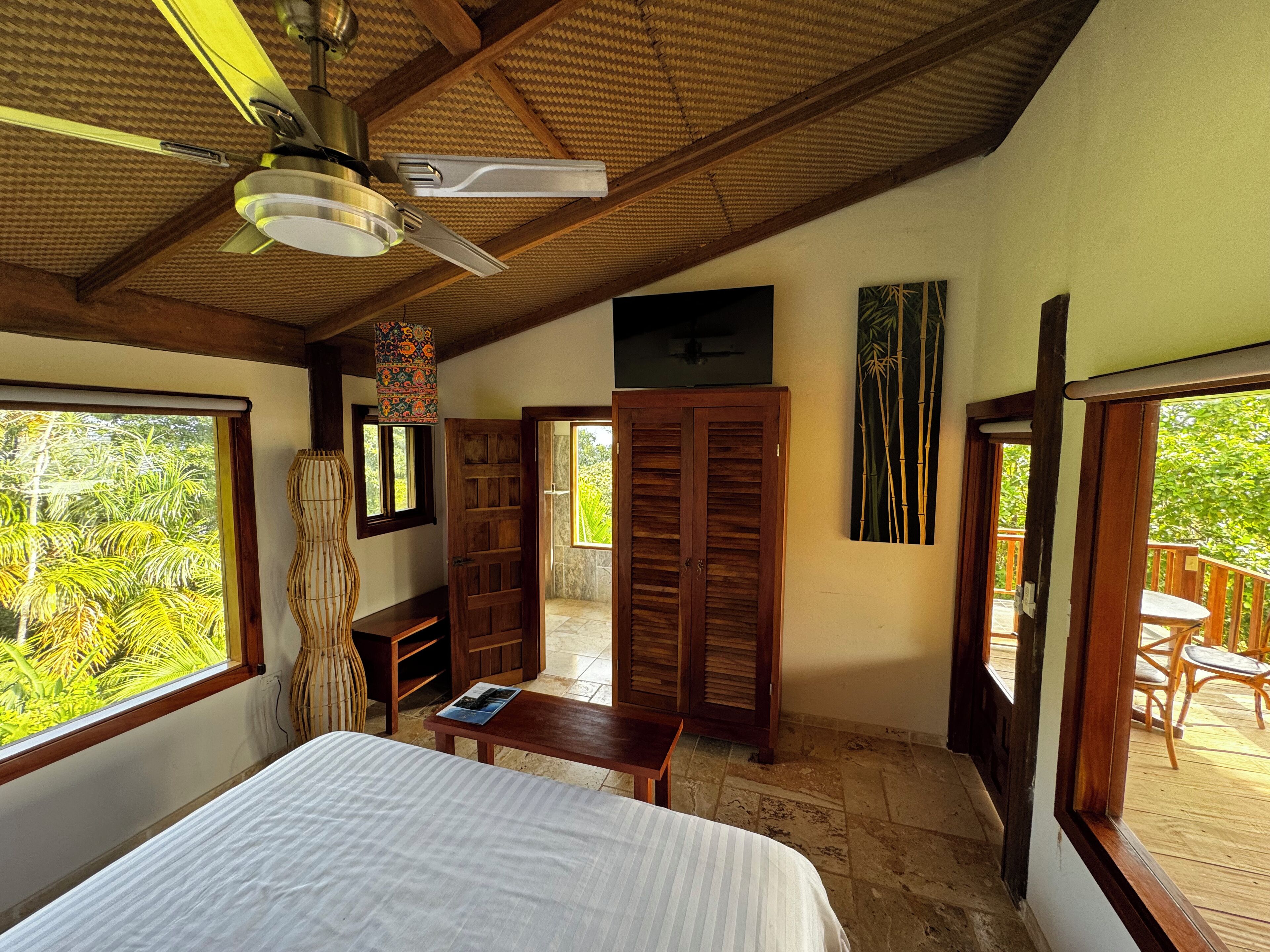 Un dormitorio con cama, ventilador de techo, ventana con vista a follaje y puerta de madera que da a otra habitación.