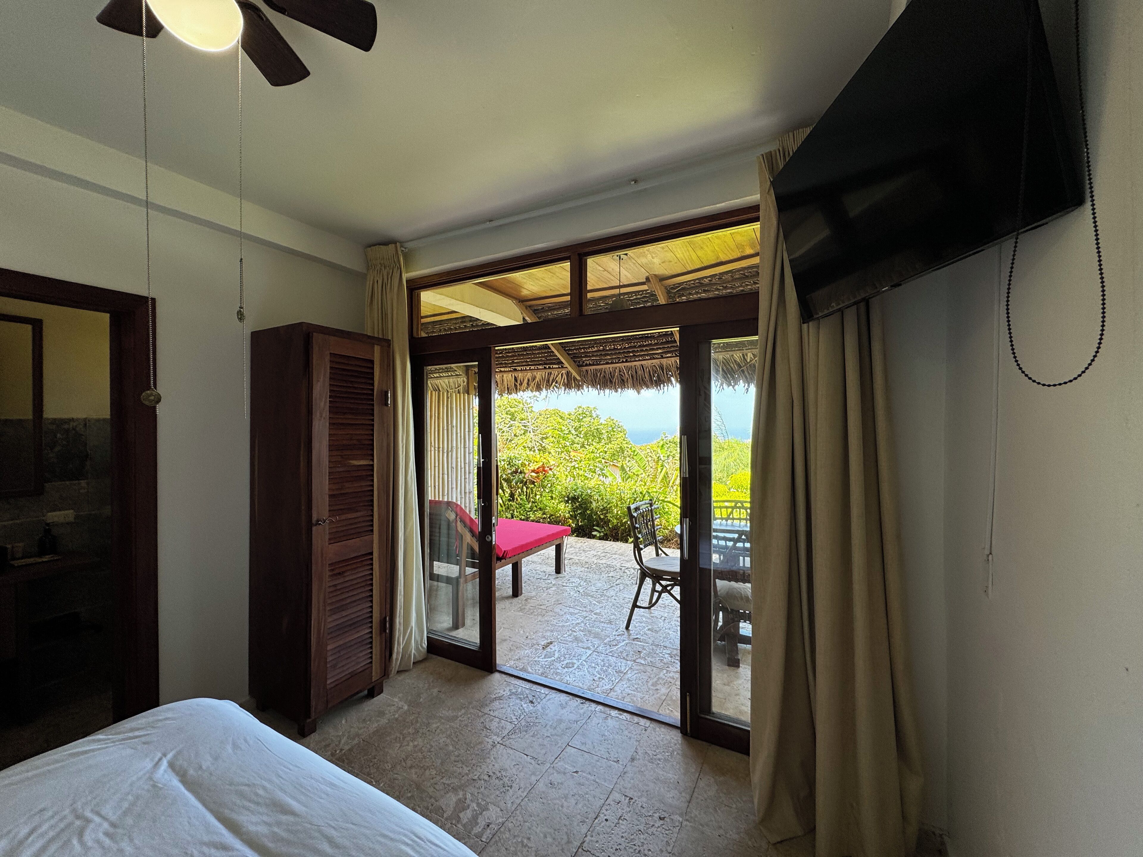 A hotel room with a bed, a TV, a wardrobe, and a sliding glass door leading to an outdoor area with a table and chairs.