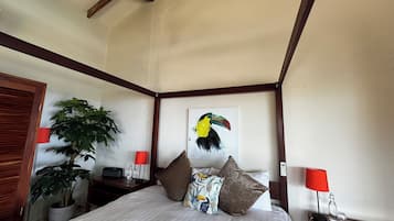 Un dormitorio con una cama grande, cabecera de madera, dos mesitas de noche, una mesita auxiliar, una lámpara roja, una botella de vidrio, un control remoto y una planta en maceta.