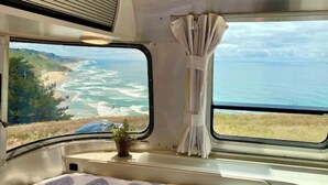 Interior de autocaravana con cama, ventana que da a vistas del océano y cortina.