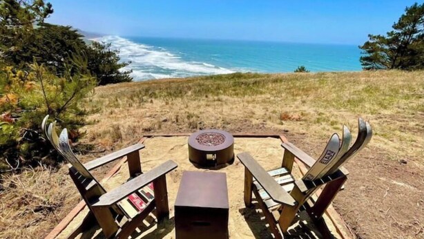Una terraza de madera con dos sillas y una mesita, con vistas a una playa y olas bajo cielos despejados.