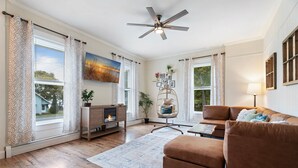 Una sala de estar con sofá marrón, chimenea, ventilador de techo y ventanas amplias con cortinas.