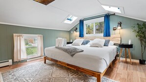 Un dormitorio con una cama grande, tragaluces, una ventana con cortinas, una mesita de noche y una planta en maceta.