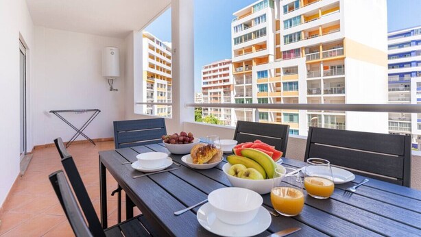 Un balcon avec une table préparée pour le petit-déjeuner, comprenant des fruits, un bol de raisins, une assiette de crêpes et un verre de jus d’orange.