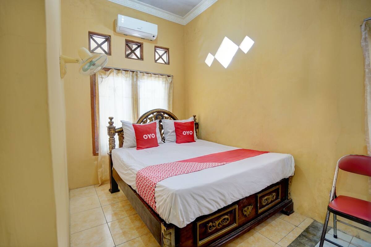 Une chambre à coucher avec un lit en bois, deux fenêtres avec des rideaux, un ventilateur de plafond et une chaise rouge.