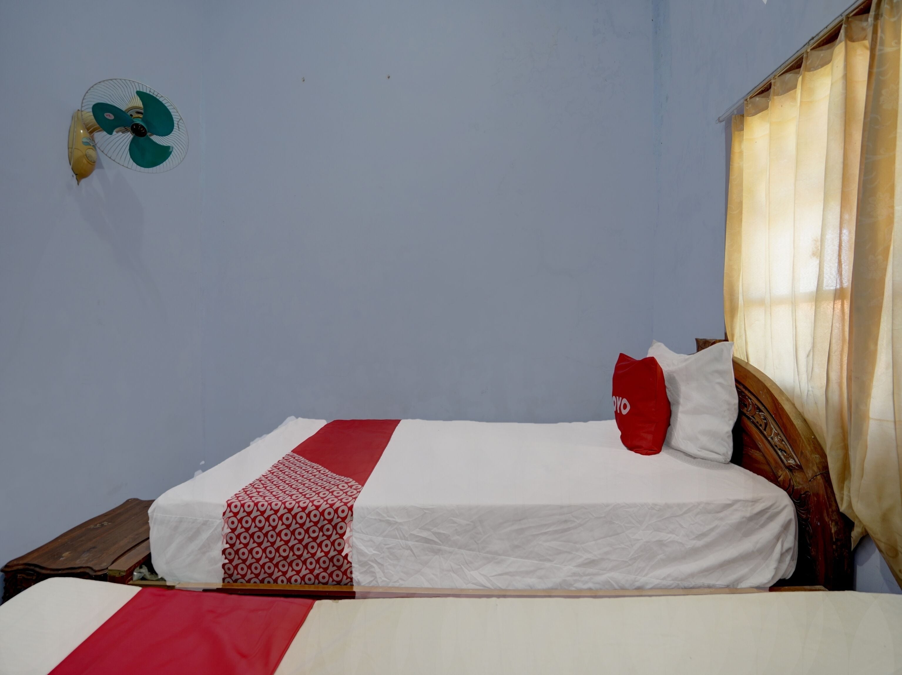 Un lit avec une couvre-lit blanche et rouge, un oreiller rouge et un ventilateur fixé au mur.