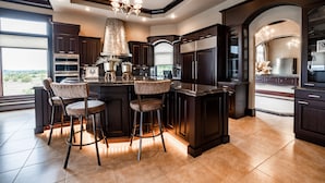 A modern kitchen with dark cabinetry, a central island with bar stools, and a large window providing a view of the outdoors. - Valley View Villa - Event Venue/Pheasant Hunting/Sleeps 18/9 Beds/6.5 Bath (Mitchell)