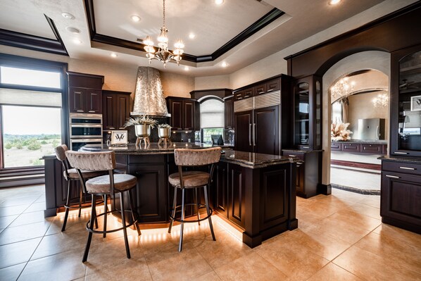 A modern kitchen with dark cabinetry, a central island with bar stools, and a large window providing a view of the outdoors.