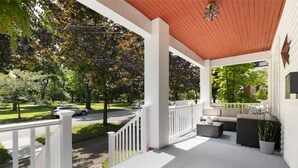 A covered porch with white railings, outdoor furniture, and a view of a street with cars and trees.