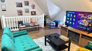 A cozy living room with a teal sofa, a black coffee table, and a TV displaying a game. There are various board games on a table, and a white crib in the background.