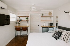 A bedroom with a bed, a desk with two chairs, a microwave, a ceiling fan, and a TV mounted on the wall. - Studio 3 at Beer Ranch Project. Walk to Wimberley Square. Unwind. Stay awhile. (Wimberley)
