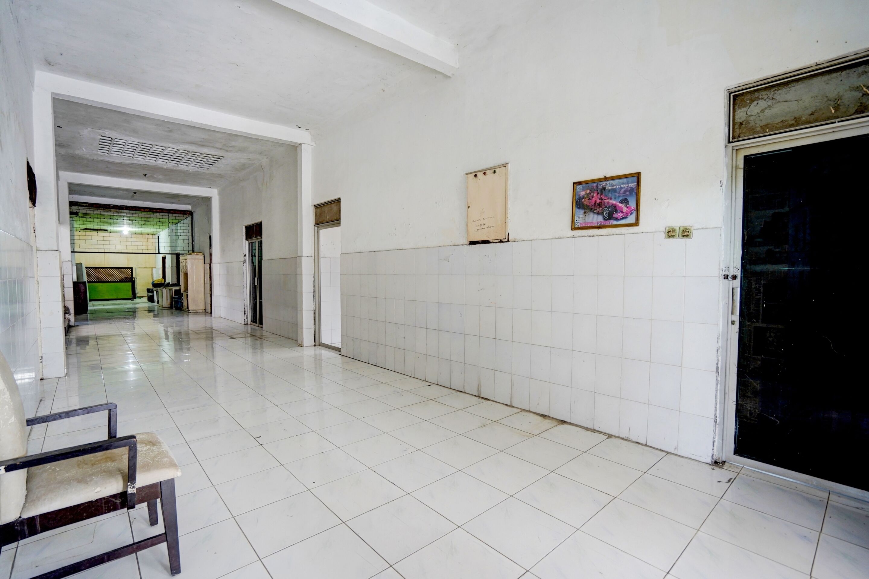 A hallway with white tiled floors and walls, a door with a black frame, and a chair in the foreground.