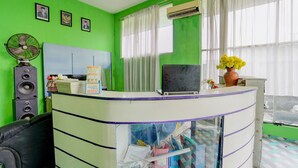 Un mostrador de recepción con una computadora portátil, un ventilador y un florero con flores. En la pared hay cuadros enmarcados y una ventana con cortinas.