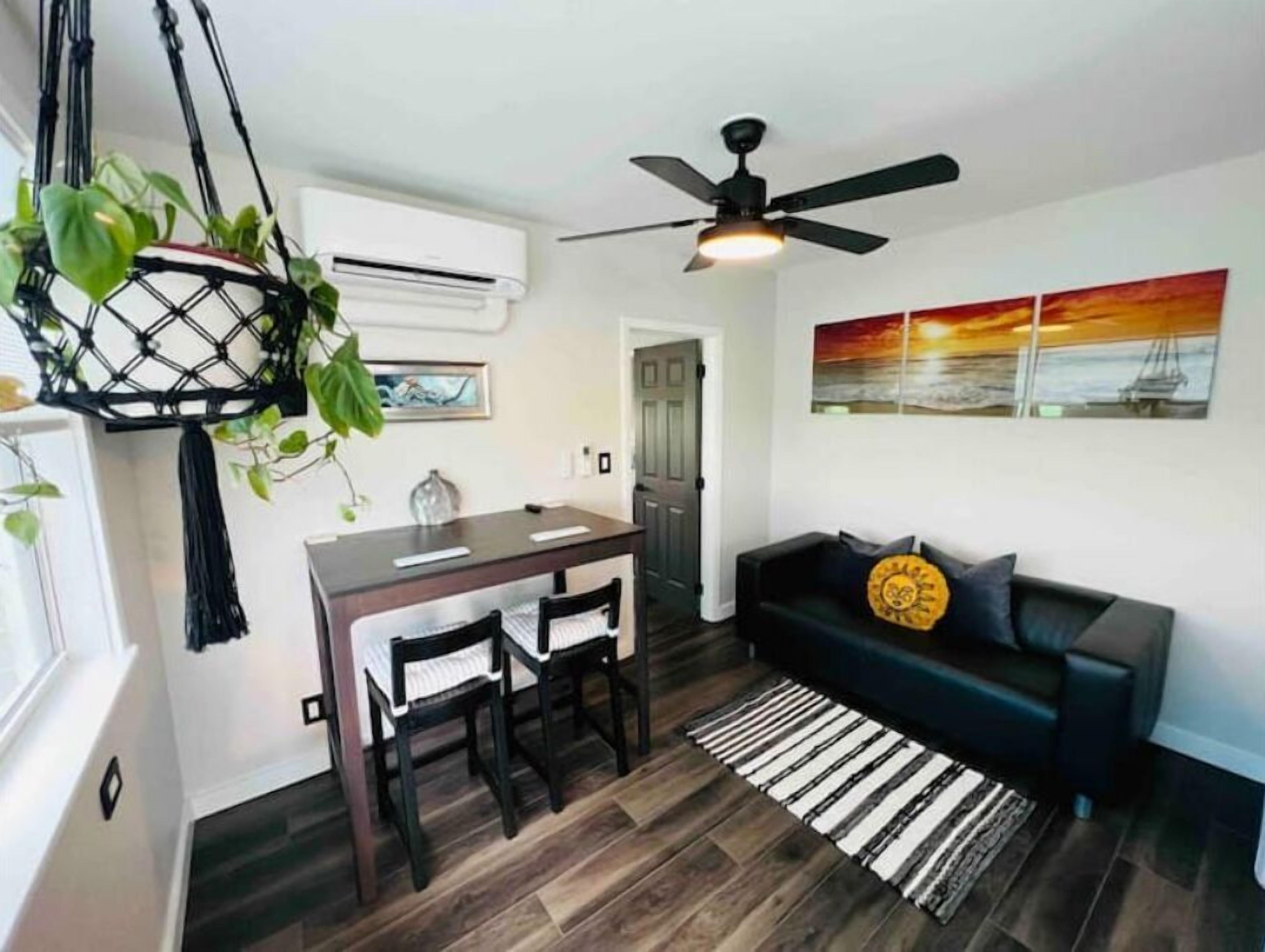Un espace de vie compact comprenant un coin repas, un canapé noir, un ventilateur de plafond et un climatiseur fixé au mur.