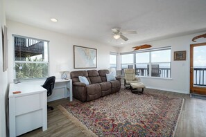 A spacious living room with a large window, a ceiling fan, a brown sofa, a white desk, and a colorful rug.