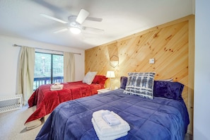 A bedroom with two beds, a ceiling fan, a window with curtains, and a wooden wall panel.