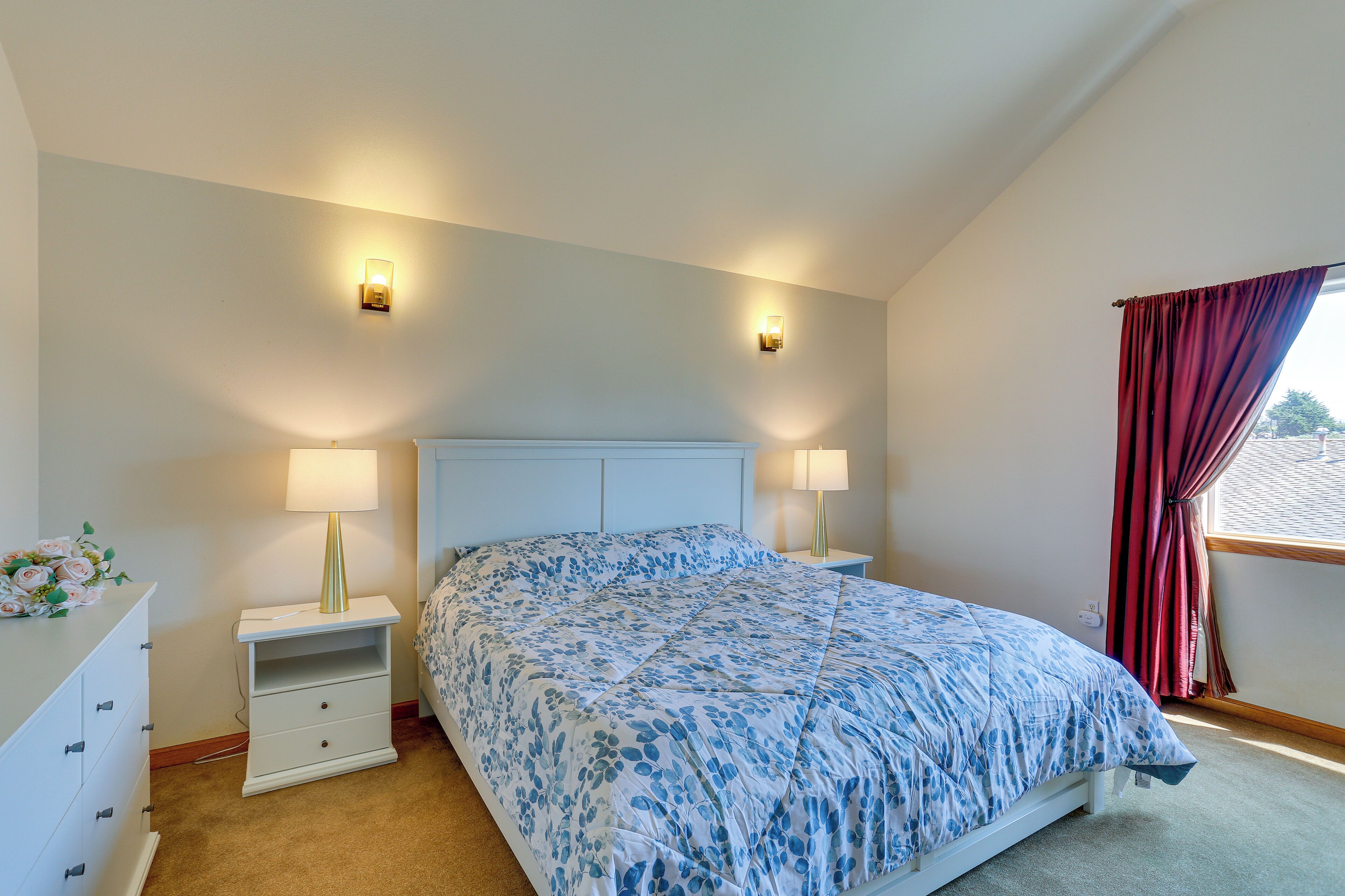 Un dormitorio con cama, dos lámparas de noche, una mesita de noche, un aparador y una ventana con cortinas.