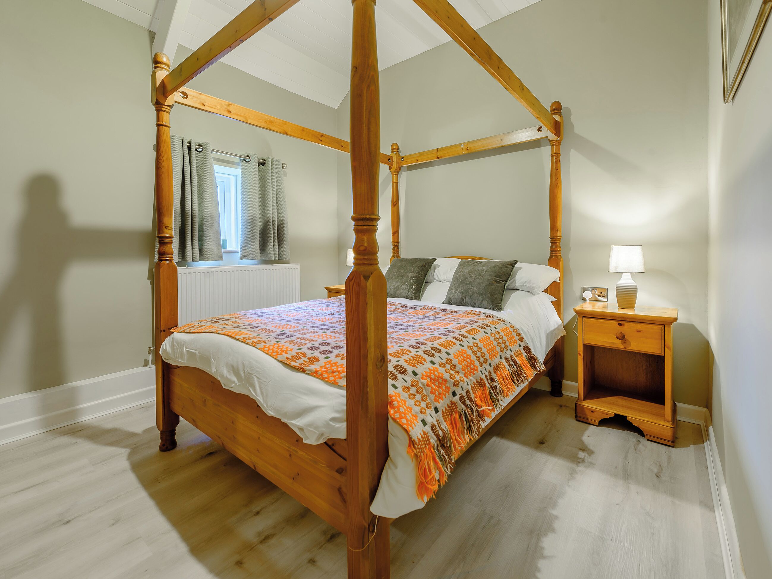 A four-poster bed with a patterned bedspread, a bedside table with a lamp, and a window with curtains.