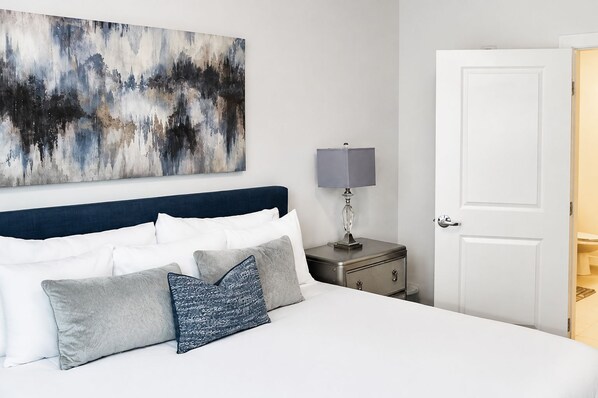 A hotel room with a bed, a nightstand with a lamp, a door, and a large abstract painting on the wall. - The Luxe | City View | Romantic Escape (Atlanta)