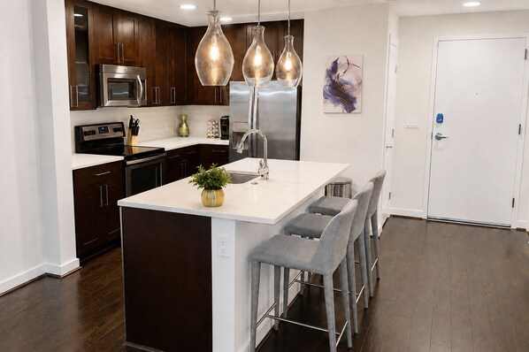A modern kitchen with dark wood cabinets, stainless steel appliances, a white island with bar stools, and pendant lighting. - The Luxe | City View | Romantic Escape (Atlanta)