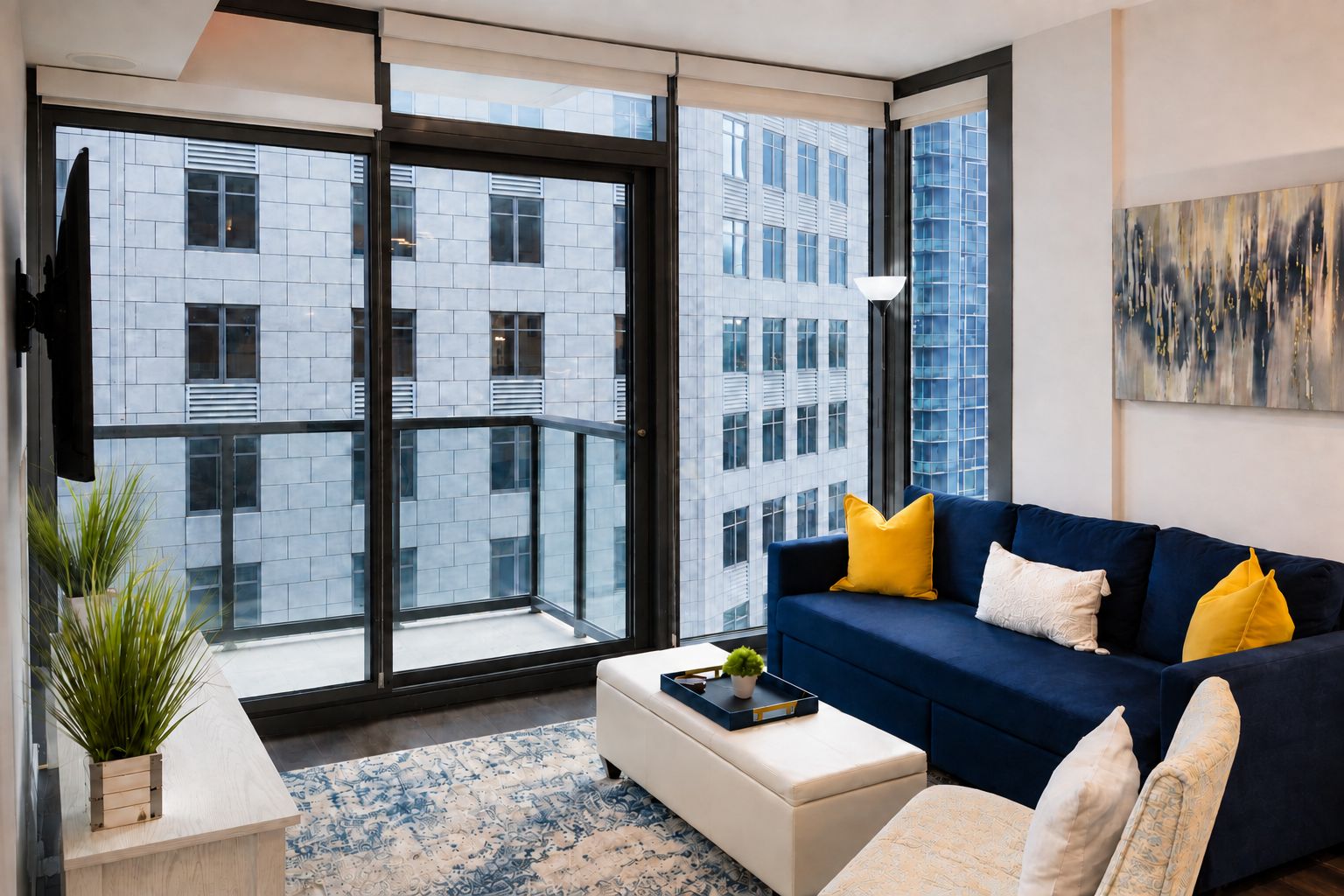 A modern living room with a blue sofa, white cushions, a coffee table, and a large window offering a view of a cityscape.